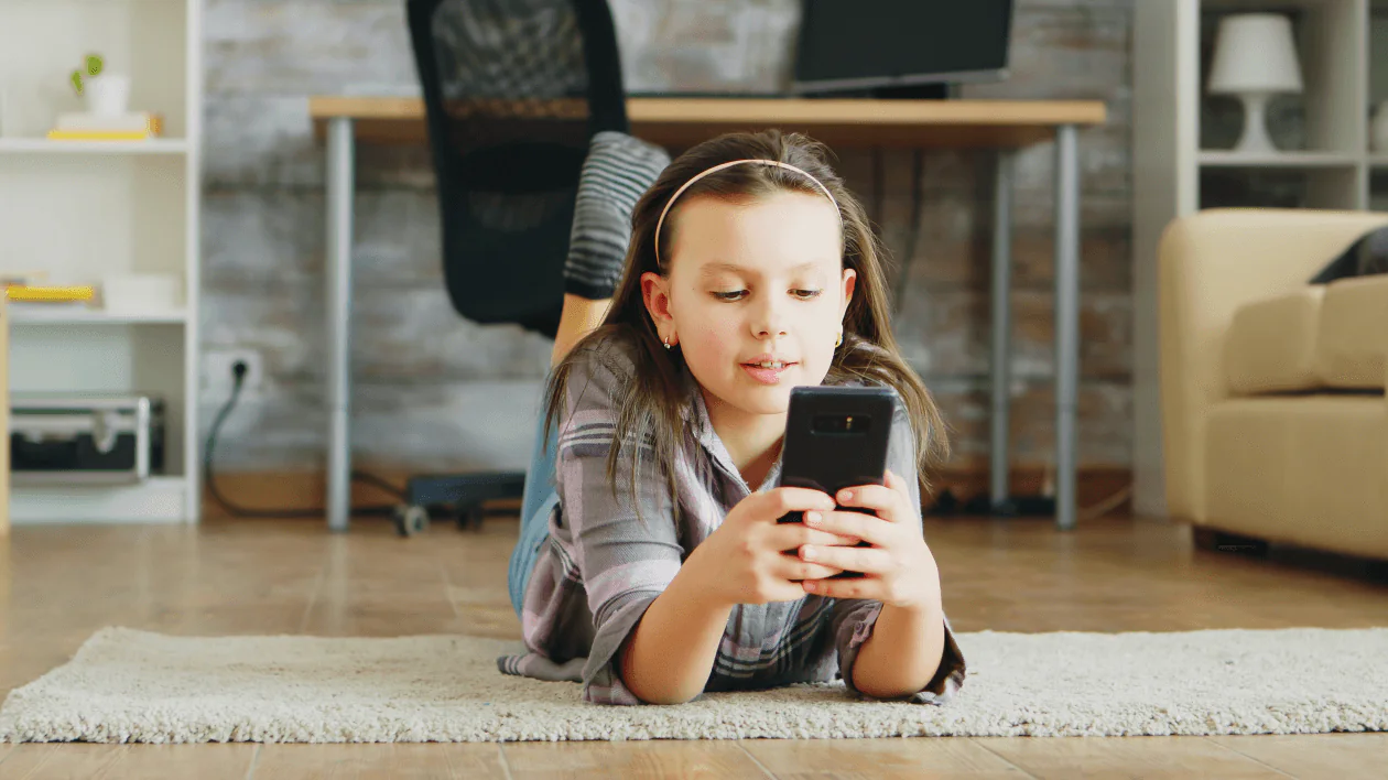 Ung flicka som ligger på en matta hemma med en mobil, som exempel på barns skärmtid utanför skolan